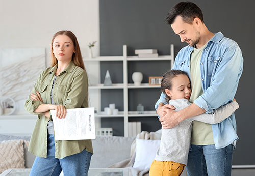 A solemn woman with papers stands apart from a father hugging his child.